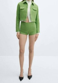 Green textured cropped jacket with matching shorts, paired with black pointed-toe heels. Features two front pockets and a zipper.