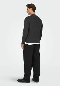 Maglione grigio scuro con collo tondo, polsini a coste e vestibilità comoda, indossato su una camicia bianca, abbinato a pantaloni neri larghi e scarpe.