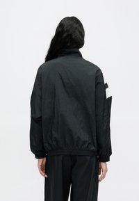 Svart, lett jakke med ståkrage og elastiske mansjetter. Har hvite geometriske aksenter på ermene. Glatt, teksturert stoff.