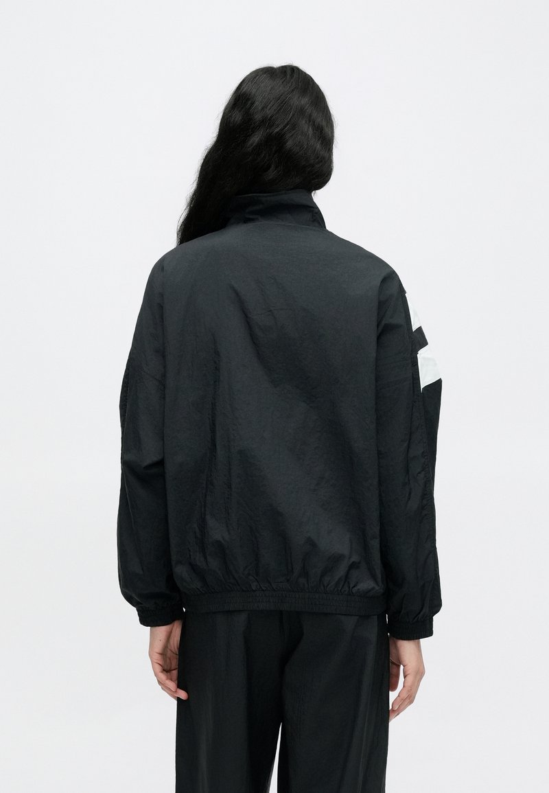 Sort, let jakke med opretstående krave og elastiske manchetter. Har hvide geometriske accenter på ærmerne. Glat, tekstureret stof.