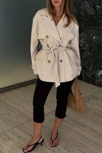 Bežinis dviem eilėmis sagų paltas su juostele, didelėmis saga, ir kišenėmis, derinamas su juodomis trumpomis kelnėmis ir juodais šlepetėmis.