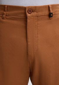 Braune Baumwollhose mit strukturiertem Finish, ausgestattet mit einem Knopfverschluss und einem kleinen schwarzen Akzentlogo in der Nähe des Bundes.