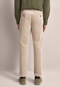 Beige Cordhosen mit geradem Schnitt, umgeschlagenen Bündchen und zwei Gesäßtaschen. Getragen mit einem grünen Hemd und grauen Slippers.