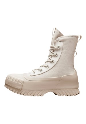 Bottes montantes blanches avec une tige en toile, un capuchon en caoutchouc, une semelle texturée et des œillets en métal avec des lacets. Boucles latérales pour un enfilage facile.