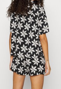 Osoba w czarnej koszulce z krótkim rękawem i szortach z białym abstrakcyjnym kwiatowym wzorem, pokazana z tyłu na jednolitym tle.