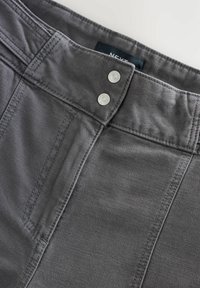 Grå denimbyxor har en platt midja med två silverknappar, synlig sömnad och en etikett inuti som visar varumärket.