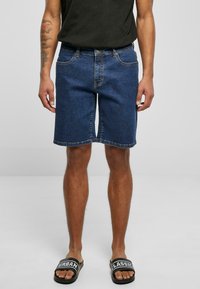 Shorts en denim bleu foncé avec coupe droite, cinq poches et couture contrastée. Portés avec une chemise noire et des sandales noires.