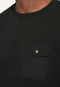 Svart t-shirt med kort ärm, tillverkad av bomull, med en bröstficka med knapp, rund hals och slät yta.