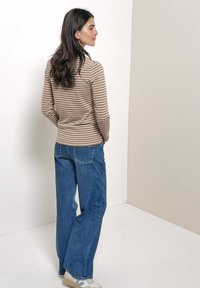 Femme aux longs cheveux foncés portant un haut beige et blanc rayé à manches longues, un jean bleu à jambes larges et des baskets blanches, debout de côté.
