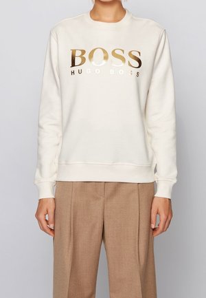 Osoba ubrana w białą bluzę z błyszczącym złotym tekstem "BOSS HUGO BOSS" i jasnobrązowe spodnie na jednolitym szarym tle.