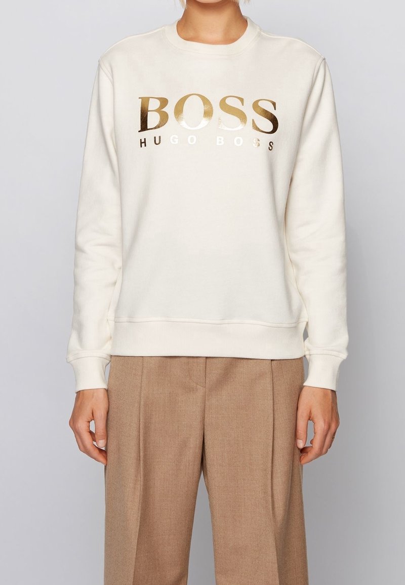 Personne portant un sweat-shirt blanc avec un texte "BOSS HUGO BOSS" doré brillant et un pantalon marron clair sur un fond gris uni.