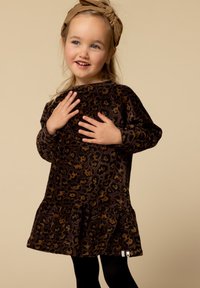 Lachend meisje in een bruine jurk met luipaardprint en bijpassend haarbandje, met haar handen voorzichtig op haar borst geplaatst.