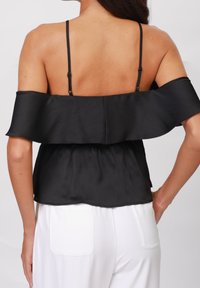 Haut noir décolleté épaules avec un design à volants, bretelles fines, texture côtelée et taille froncée, associé à un pantalon blanc.