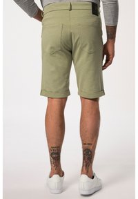 Pantaloni corti verde chiaro con orli arrotolati, tasche posteriori e un'etichetta cucita in vita. Indossati con scarpe da ginnastica bianche. Tatuaggi visibili sulle gambe inferiori.
