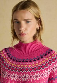 Svetlo roza pletena jopica z visokim rebrastim ovratnikom in barvnimi vzorci, tematsko povezani z zimskim časom, ki vključujejo geometrijske oblike in cvetlične akcenta.