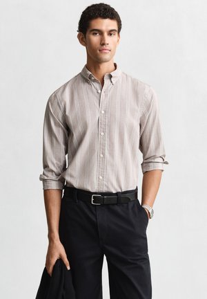 Junger Mann trägt hell gestreiftes Hemd mit aufgekrempelten Ärmeln, schwarze Hose, schwarzen Gürtel und silberne Armbanduhr, hält schwarze Jacke.