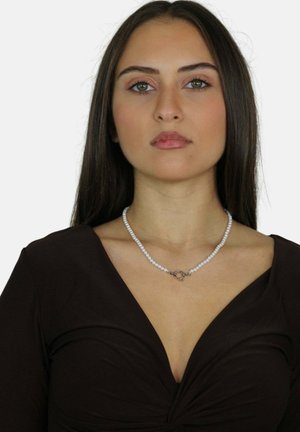 Collana di perle con chiusura in argento, caratterizzata da perle bianche rotonde, indossata da una modella con un top marrone a maniche lunghe e scollo a V profondo.