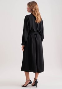 Donna con capelli ondulati che indossa un vestito midi nero a maniche lunghe, con vita cintrata, e sandali con tacco alto neri, in piedi di lato.