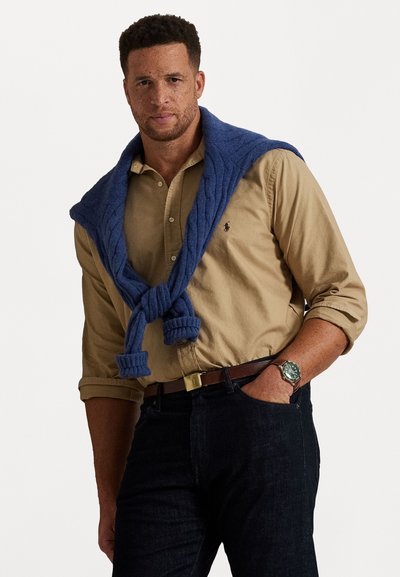 Kék kötött pulóver, amely egy világosbarna gombos ingre van terítve, sötét farmernadrággal és ezüst szíjjal párosítva. Az anyag bordázott textúrájú.