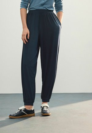 Dunkelblaue, elastische Hosen mit schmal zulaufenden Beinen, aus einem glatten Stoff. Kombiniert mit schwarzen Sneakers mit weißen Akzenten.