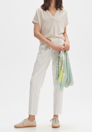 Kvinde iført cremefarvet top med V-udskæring, hvide bukser, beige sneakers, holder et sammenfoldet pastelgrønt tørklæde med mønster i begge hænder.