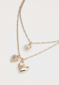 Collier superposé en ton doré composé de trois charms en forme de cœur : un poli, un avec des pierres claires, et un cœur en cristal transparent.