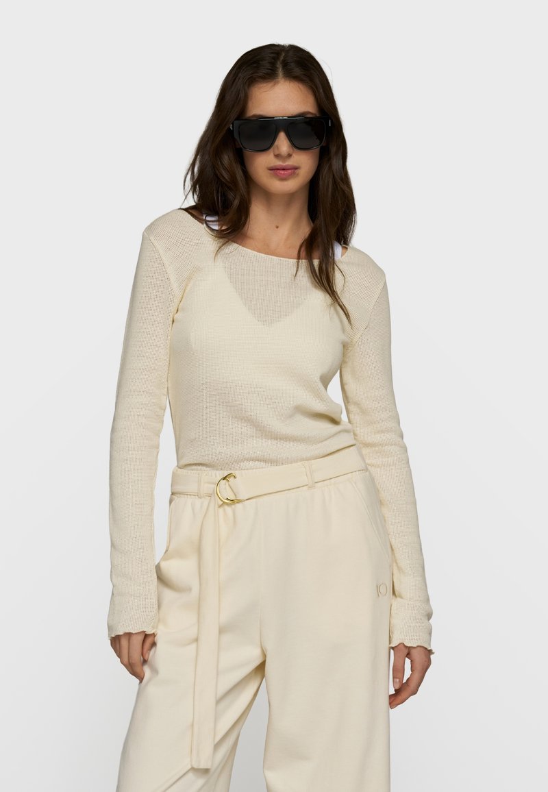 Femme portant de larges lunettes de soleil noires, un haut en maille crème à manches longues et un pantalon taille haute crème avec ceinture, sur un fond clair uni.