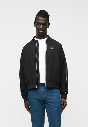 Moški v črni jakni z zadrgo in belim poudarkom čez belo majico ter modrimi kavbojkami, stoji ob svetlo sivem enobarvnem ozadju.