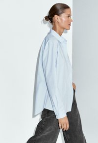 Frau mit Hochsteckfrisur trägt ein hellblaues, lockeres Hemd und schwarze Jeans, geht vor einem weißen und grauen Wandhintergrund entlang.