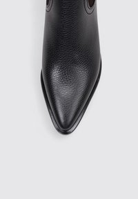 Botín de cuero negro hasta el tobillo con punta afilada, superficie texturada, costuras sutiles y tacón redondeado, situado sobre un fondo gris claro.