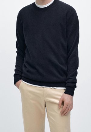Mand iført sort langærmet sweater over hvid skjorte og beige bukser, stående med den ene hånd i lommen mod en ensfarvet baggrund.