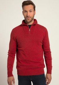 Pull rouge à col zippé avec col et poignets côtelés. Texture douce, design ajusté. Porté sur une chemise noire et associé à un jean foncé.
