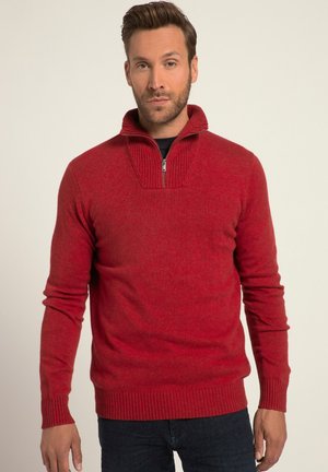 Pull rouge à col zippé avec col et poignets côtelés. Texture douce, design ajusté. Porté sur une chemise noire et associé à un jean foncé.