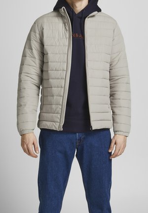 Mann trägt beige gesteppte Jacke über einem dunkelblauen Hoodie und blauen Jeans, steht vor einfachem grauem Hintergrund.