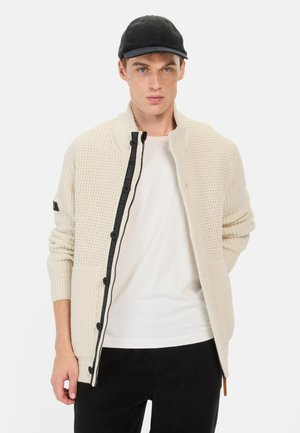Creme strikket cardigan med struktureret mønster, sort lynlås og knapper, to forlommer, båret over en hvid skjorte, parret med en sort kasket.
