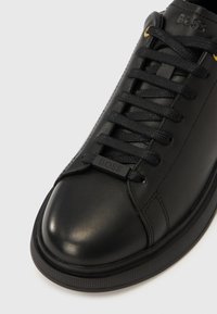 Schwarzer Ledersneaker mit schwarzen Schnürsenkeln, goldfarbenen Ösen und "BOSS"-Logo auf Zunge und Schnürsenkelanhänger, von oben auf weißem Hintergrund gezeigt.