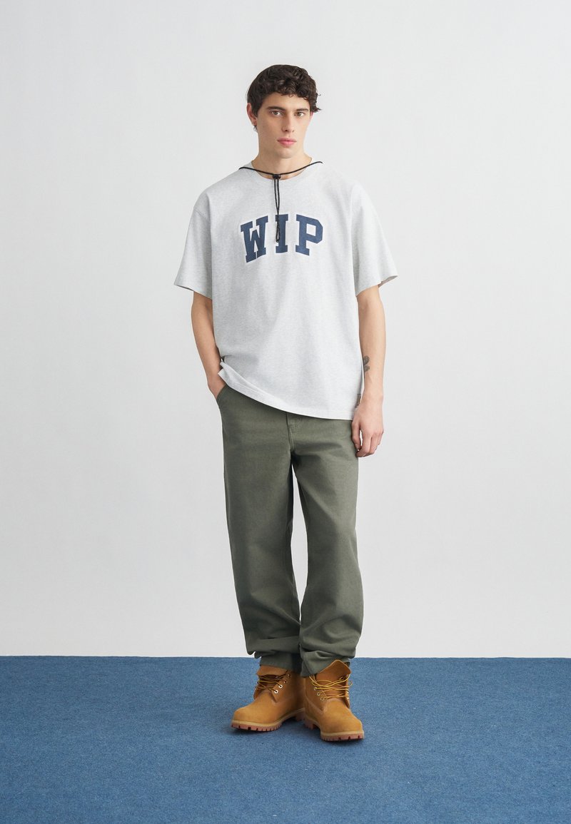 Junger Mann steht drinnen und trägt ein weißes WIP-T-Shirt, olivgrüne Hosen und hellbraune Schnürstiefel auf einem blauen Teppich.