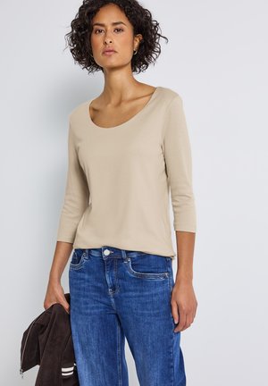 Haut beige à manches longues avec un encolure dégagée, associé à un jean en denim bleu. Le modèle tient une veste marron foncé avec des accents blancs.