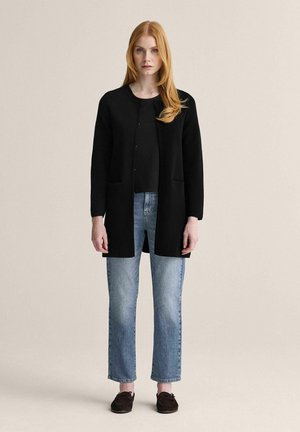 Ung kvinde med langt rødt hår iført sort åben cardigan, sort top, lyseblå jeans og mørke mokkasinsko, stående mod en beige væg.