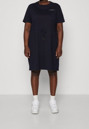 Persoon draagt een zwarte knie-lange jurk met korte mouwen, een taille met trekkoord, witte sneakers en witte sokken tegen een effen achtergrond.