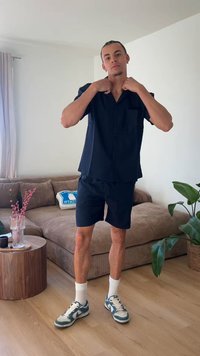 Άνδρας με μπλε σκούρο κοντομάνικο πουκάμισο και σορτς ρυθμίζει το γιακά, στέκεται σε σαλόνι με καφέ καναπέ, αθλητικά παπούτσια και φυτά.