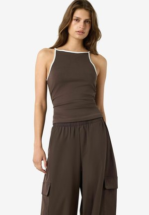 Jeune femme portant un haut sans manches marron foncé avec des bordures blanches et un pantalon cargo marron ample, debout devant un fond blanc uni.