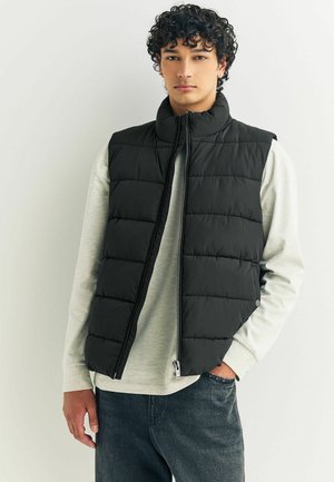 Black puffer vest with a high collar and zip closure, featuring quilted panels. Worn over a light gray long-sleeve shirt.