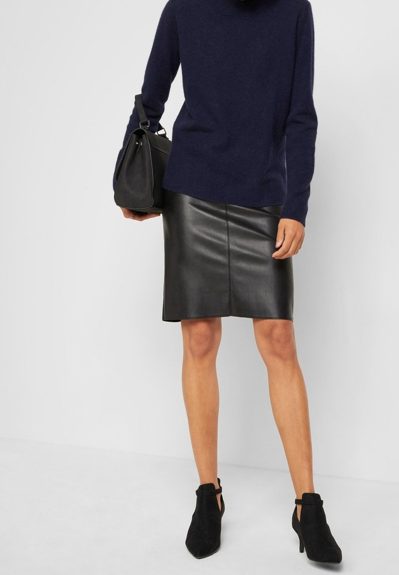 Femme portant un pull marine, une jupe en cuir noire mi-longue, des bottines noires et tenant un sac à main noir sur un fond uni.
