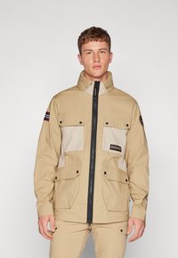 Giacca beige con chiusura a zip, tasche anteriori e pannello contrastante di un beige chiaro. Presenta un colletto alto e il logo sul petto.
