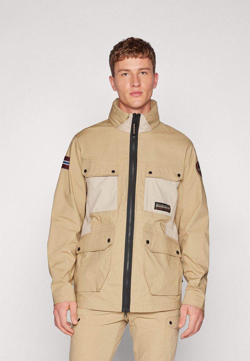 Giacca beige con chiusura a zip, tasche anteriori e pannello contrastante di un beige chiaro. Presenta un colletto alto e il logo sul petto.