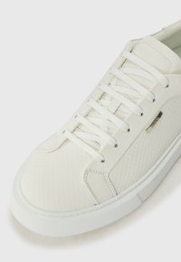 Primo piano di una sneaker bianca con lacci, caratterizzata da un motivo esagonale testurizzato e un dettaglio in metallo sul pannello laterale.