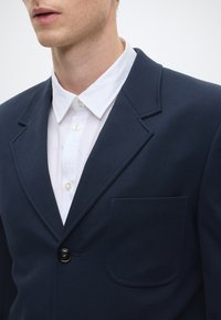 Blazer azul-marinho com fecho de botão único, lapela notched e um bolso de remendo, sobre uma camisa branca de colarinho. Textura de tecido lisa.