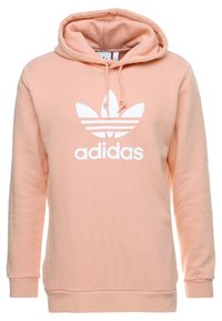Persikofärgad Adidas hoodie med en framficka i kängurustil, dragsko i huvan och en stor vit logotyp med tre blad och varumärkesnamn.