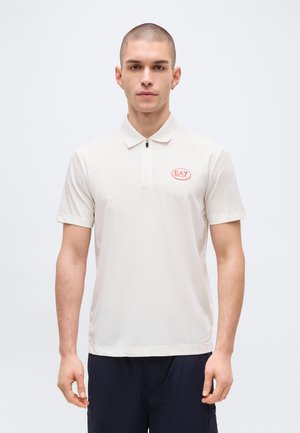 Noor mees lühikeste juustega, valges EA7 polo-särgis ja tumedates pükstes, seisab ühevärvilise heleda tausta ees.